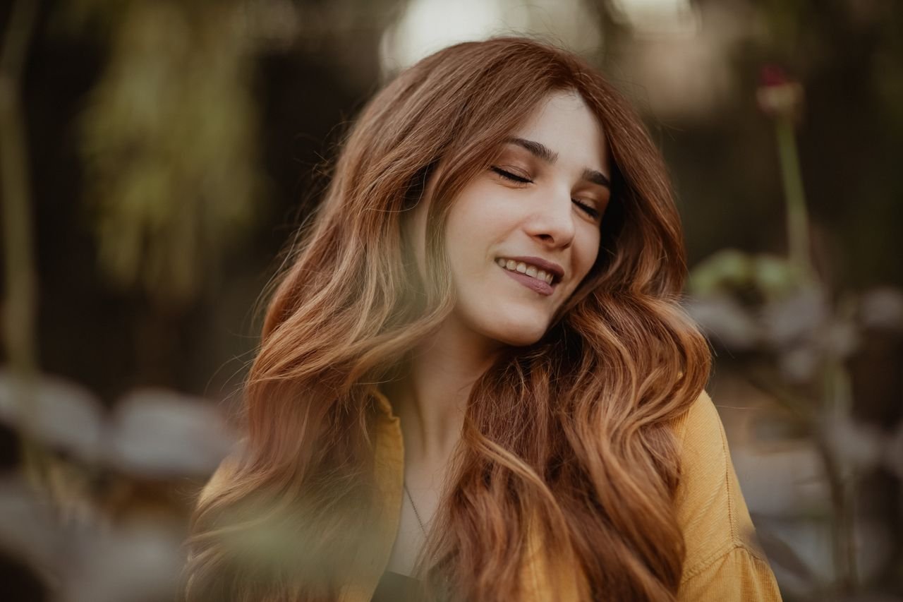 Portrait of a smiling woman with long red hair and eyes closed, outdoors in a natural setting.